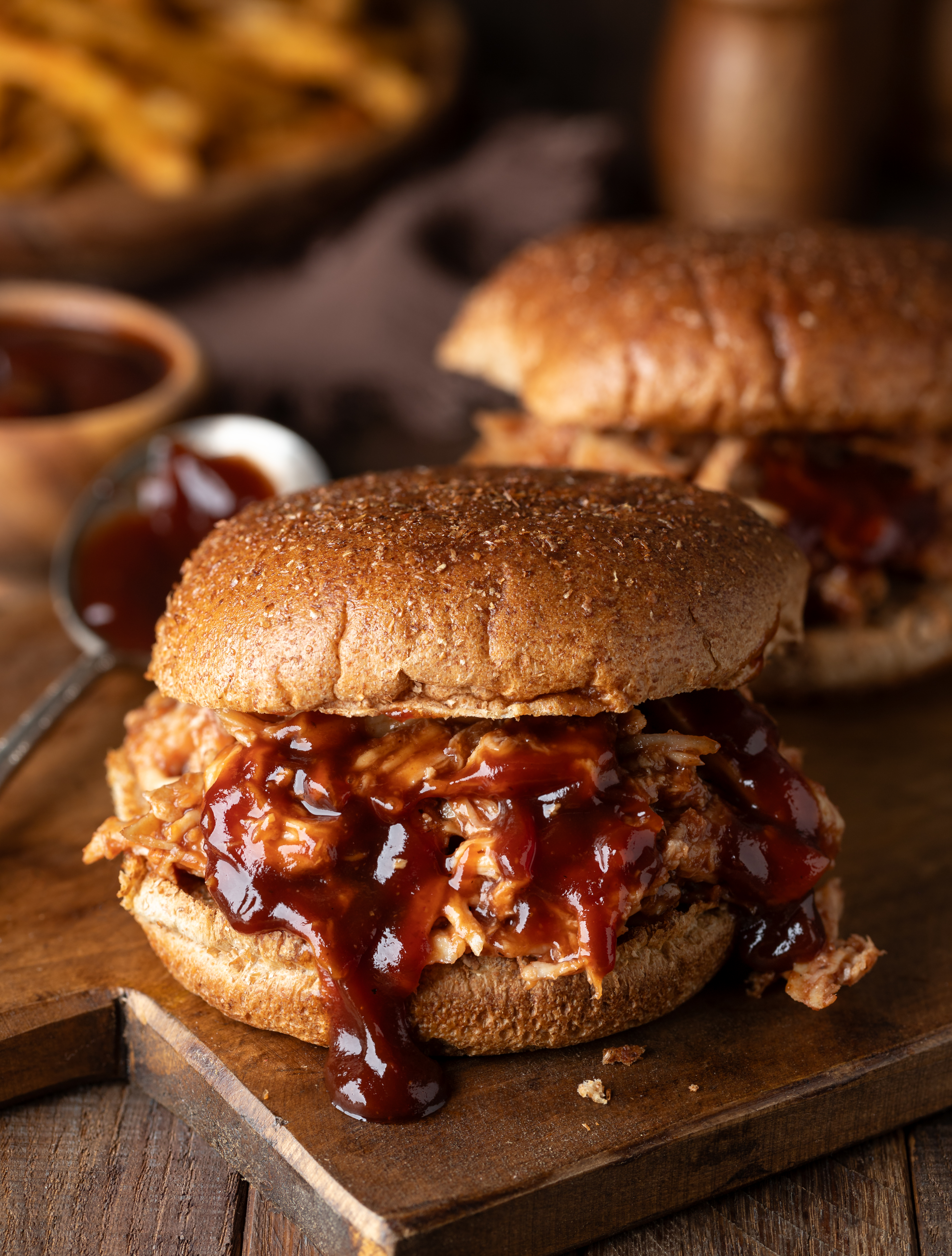 Barbeque pulled chicken sandwichs on whole wheat bun on wooden cutting board with french fries in background