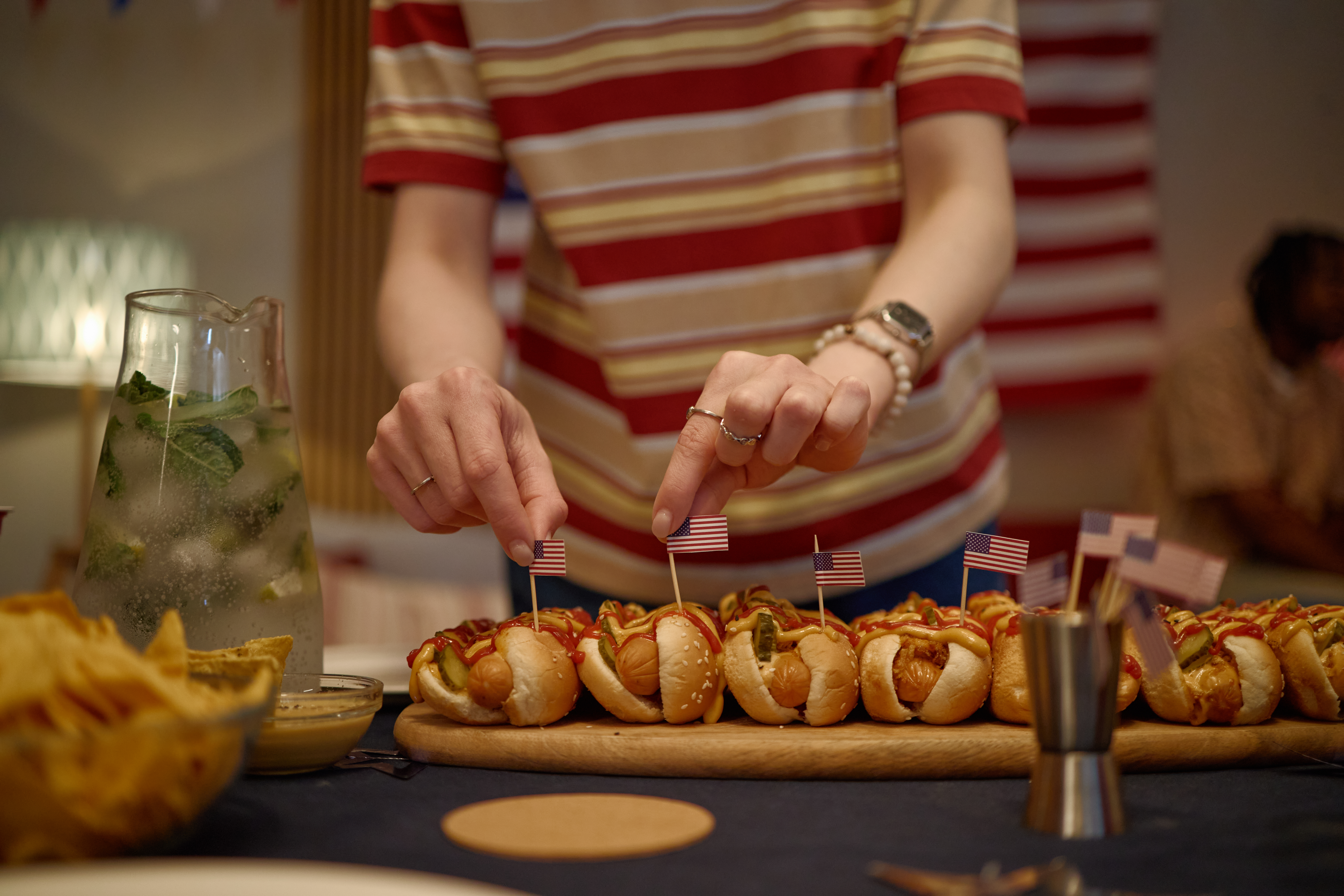 Young adult woman arranging mini American flags on hot dogs during home Independence Day gathering, showing patriotic entertaining. Ideal for holiday marketing, party promotion