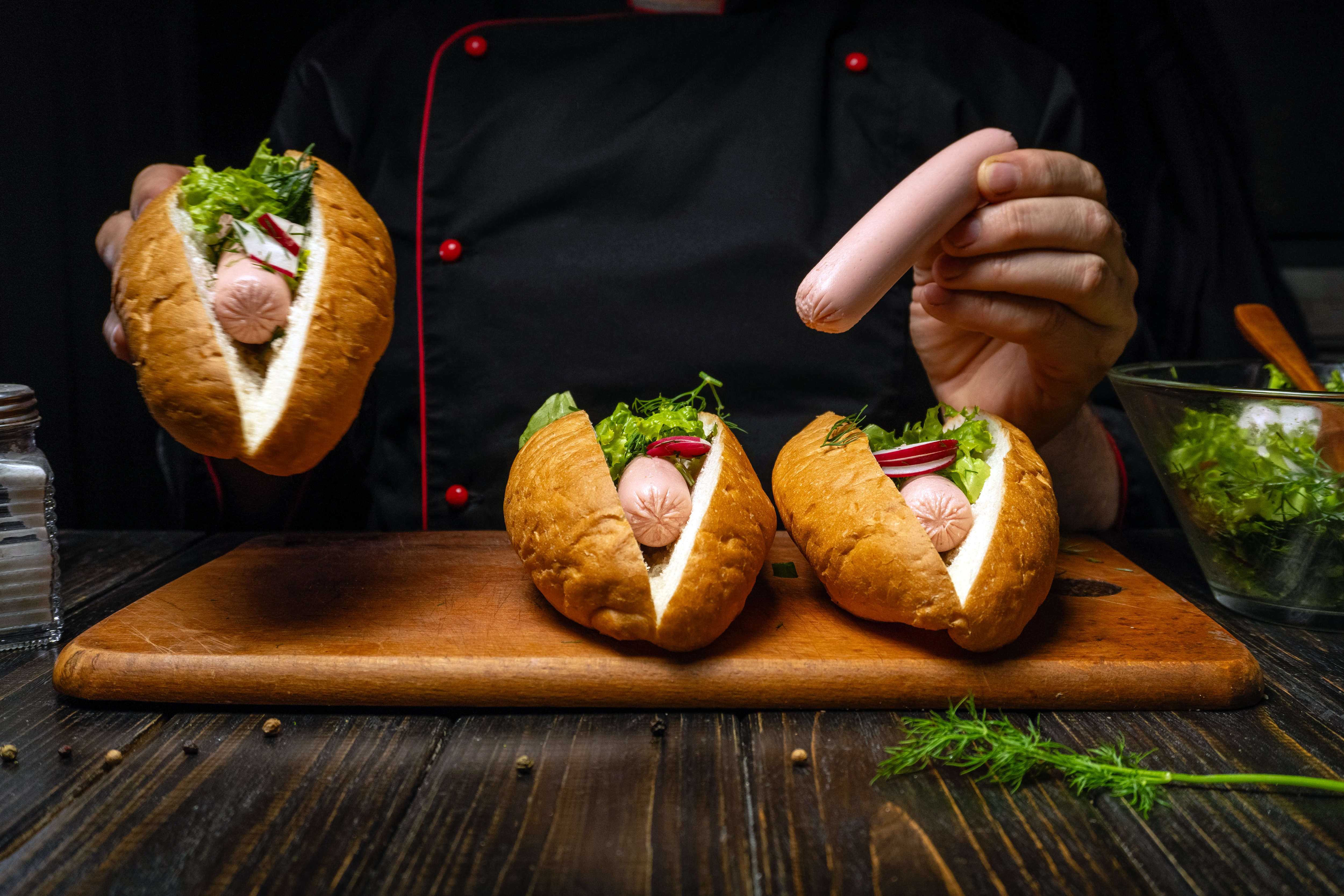 A chef holds hot dogs filled with vegetables. Three hot dogs are placed on a wooden board. The chef is working in a kitchen in the evening. A bowl of salad is nearby.